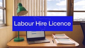 Office desk with laptop and documents beside a green lamp, featuring a blue “Labour Hire Licence” banner and Victoria outline overlay.